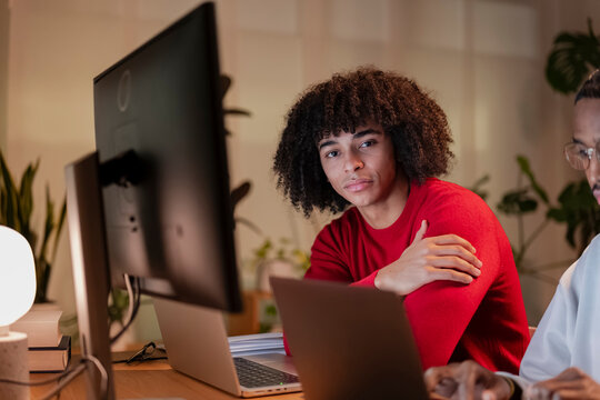 Two young people work on laptops at a desk in a dimly lit room, one looking directly at the camera.