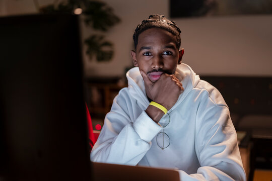 A young man with braided hair wearing a white hoodie sits at a desk, looking thoughtfully at a computer screen in a dimly lit room.