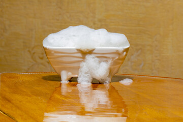 A white ceramic sponge holder shaped like a miniature bathtub, overflowing with soap foam that spills onto a polished wooden surface against a wood-grain backdrop.