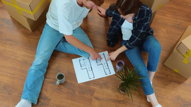 Cheerful man and woman sitting on the floor in new house with coffee and home project discussing interior design and reconstruction. Family moving in new home. Top view. Slow motion
