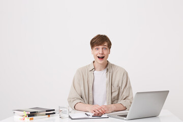 Closeup of surprised young man student with braces wears beige shirt feels impressed and using laptop computer and notebooks at the table isolated over white background