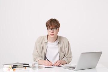 Closeup of serious handsome young man student wears beige shirt and glasses sitting with laptop computer and notebooks at the table and writing isolated over white background