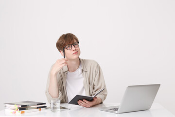 Closeup of pensive handsone young man student wears beige shirt thinking and writing in notebook at the table with laptop computer isolated over white background