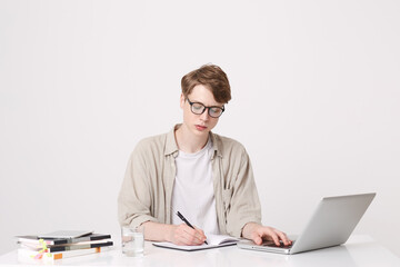 Portrait of serious young man student wears beige shirt and spectacles writing and study at the table using laptop computer and notebooks isolated over white background