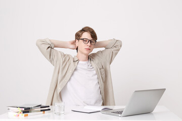 Closeup of relaxed confident young man student wears beige shirt and glasses sitting with hands over head at the table with laptop computer and notebooks isolated over white background