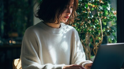 窓辺でパソコン作業に集中する女性の姿。仕事や学習シーンに適したイメージ素材。
