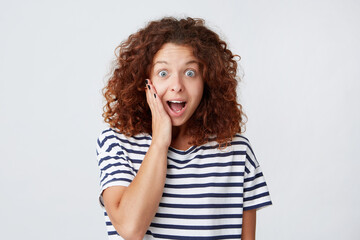 Portrait of surprised excited cute young woman with curly hair and opened mouth wears striped t shirt looks amazed and shouting isolated over white background