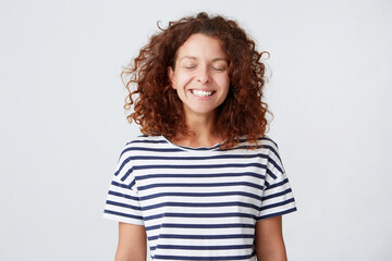 Closeup of cheerful pretty young woman with curly hair wears striped t shirt keeps eyes closed and making a wish isolated over white background Looks happy