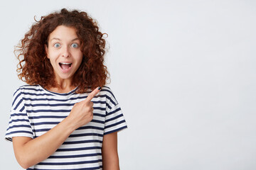Portrait of amazed happy young woman with curly hair an opened mouth wears striped t shirt looks surprised and points to the side isolated over white background
