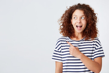 Closeup of surprised beautiful young woman with curly hair and opened mouth wears striped t shirt looks and points to the side isolated over white background Feels happy