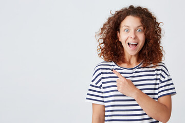 Cheerful beautiful young woman with curly hair wears striped t shirt feels excited and points to the side isolated over white background