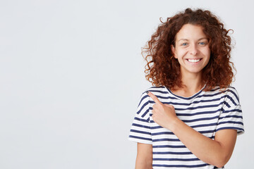 Portrait of happy attractive young woman with curly hair wears striped t shirt stands and points to the side with finger isolated over white background