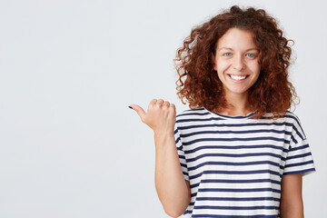 Closeup of smiling pretty curly young woman wears striped t shirt looks confident and points to the side isolated over white wall Feels happy
