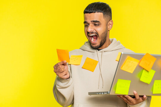 Exhausted freelancer Indian man with pasted sticker notes use laptop computer having concentration problem creative crisis. Nervous Arabian guy confused by big routine work loading yellow background