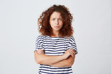 Closeup of serious worried young woman with curly hair wears striped t shirt feels embarrassed and stands with arms crossed isolated over white background