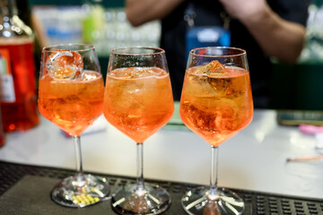 Three stemmed wine glasses filled with orange Aperol Spritz cocktails and ice cubes rest on a bar counter