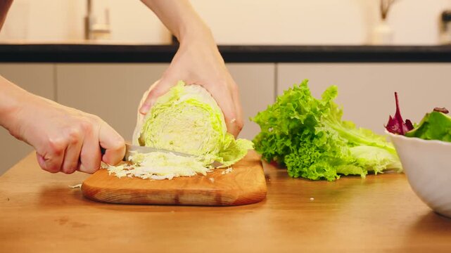 Close-up shot person hands with knife cutting Chinese naba cabbage on wooden board for healthy food vegetable diet dinner. woman routine preparing vegan fresh green salad on table white kitchen home