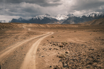 Winding dirt road cutting through a rugged, arid mountain canyon landscape under a dramatic sky. A remote path leading into the wilderness