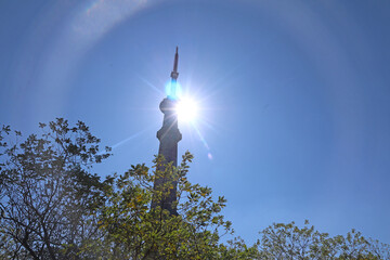 Mhaismal, MH, India, Dec 12: Low angle view of the Doordarshan tower, silhouetted by the sun, which creates a strong starburst effect and lens flare against a clear blue sky, framed by trees.