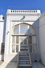 Khuldabad, MH, India, Dec 12: Detailed white marble entrance with a scalloped arch, geometric jali screens, steps, and a white wall topped with modern razor wire.