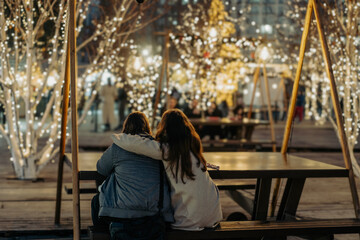 A hugging couple sitting at a Christmas market decorated with garland, shot from the back