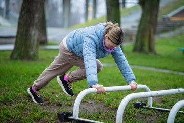 A woman in a blue puffer jacket and athletic wear exercises on outdoor equipment in a park, performing a plank or push-up style movement. She is focused on her fitness routine.
