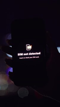 Vertical video. Smartphone Displaying SIM Not Detected Warning Held Against Night City Bokeh in a Close Angle Dark View Showing SIM Failure Situation
