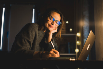 Mature happy woman working on laptop in home office at evening