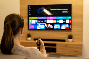 Woman watching smart TV LED in modern living room: rear view of hand holding remote control, choosing media app on screen, home entertainment concept