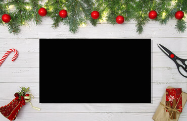 Christmas flat lay workspace with pine branches red ornaments scissors gift and black empty paper on white wooden table showing holiday crafting concept and copy space