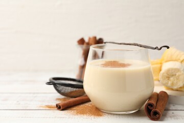 Delicious protein shake in glass, cinnamon, banana and vanilla pod on white wooden table, closeup. Space for text