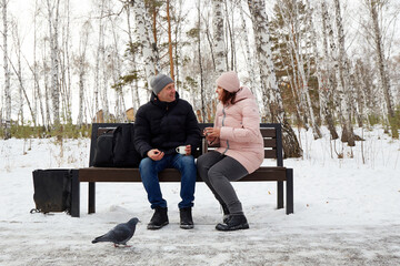  A man and woman are relaxing in a snowy winter park. They are sitting on a bench, drinking hot tea from a thermos