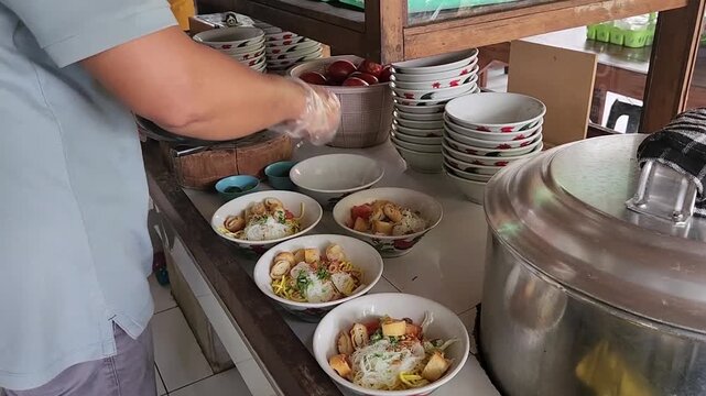 Adding crispy fried dough to Asian noodle soup bowls. Street food chef topping noodle soup with crispy cruller. Preparing Indonesian noodle soup with fried dough topping