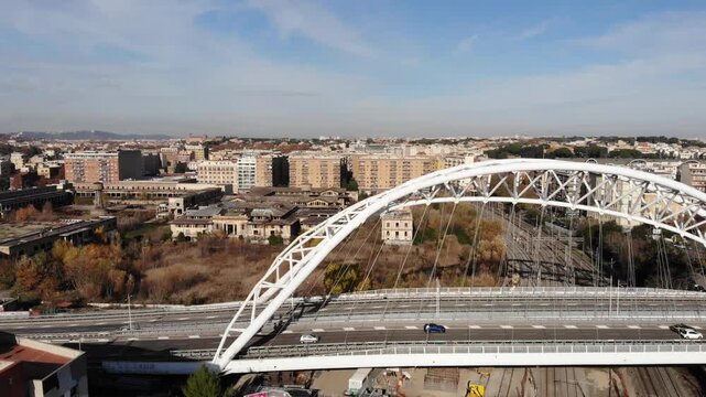 Aerial panoramic drone footage of a metal bridge, in Garbatella, Rome, with cars crossing the bridge and a train passing under and an abandoned old market in the background