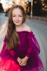 Stylish smiling princess kid girl 6-7 year old with blonde long hair wearing pink festive dress posing outdoors. Childhood.