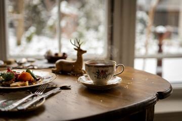 Quiet winter breakfast scene with steaming tea cup on wooden table near a window. Soft daylight, snow outside and minimal home interior create calm, cozy atmosphere.