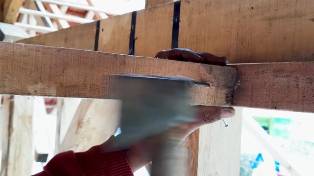 Carpenter hand driving metal nail into wooden beam structure using axe head tool during manual house construction framing work, residential building renovation and timber carpentry assembly.