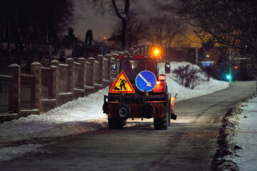 Snow removal tractor with illuminated warning lights working on icy sidewalk at night, winter maintenance. Snow plow machine with brush attachment cleaning snowy path in the street during night time