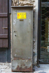 A rusted electrical cabinet with a high-voltage warning sign is mounted between old urban structures.