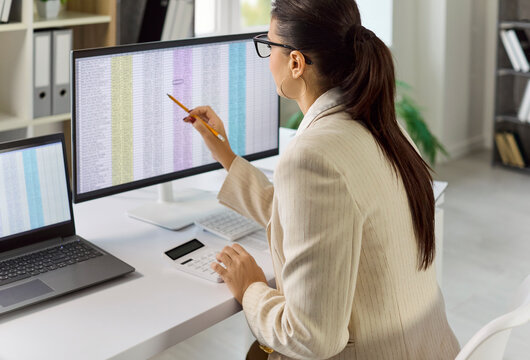 Business accountant conducts audit and analysis of data. She reviews a spreadsheet on a monitor, checks totals with calculator, and prepares reports. Theme of auditing and financial control.