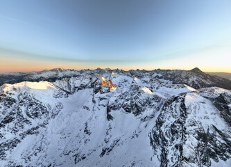 Aerial view of snow-covered mountain range during sunrise over rocky peaks in a vast landscape