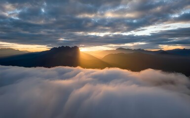 Majestic mountain sunrise above clouds, dramatic golden sun rays, aerial landscape background, foggy morning nature sky, heavenly silhouette peaks, scenic outdoor wallpaper for travel concept.
