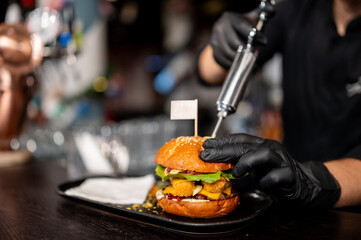 Chef in black gloves preparing a gourmet cheeseburger, injecting sauce into the bun with a syringe. Close-up on the final steps of making delicious fast food in a restaurant.