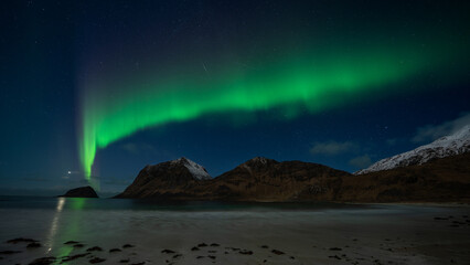 Intense northern lights dance above Haukland Beach in the Lofoten Islands, glowing over the sea and surrounding mountains in a dramatic Arctic night scene.