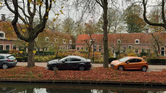 Picturesque autumn scene of a canal in a historic Dutch town. Brick houses, trees, parked cars, and calm water reflections characterize this peaceful European village landscape.

