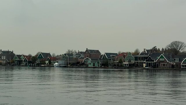 Wide panoramic panning shot across the river Zaan showcasing traditional green wooden houses and iconic historic windmills at Zaanse Schans village under a cloudy sky in the Netherlands.

