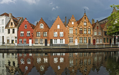 Naklejka premium Canal scene in Bruges featuring a row of traditional Flemish buildings with gabled roofs and colorful facades reflected in the canal. A dramatic sky enhances the atmosphere