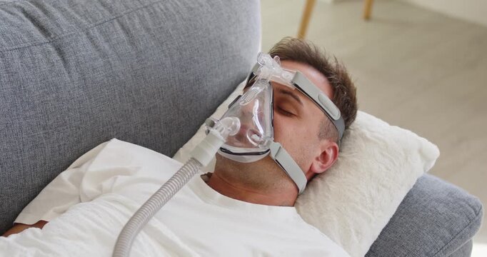Man mask sleep apnea therapy oxygen CPAP. Adult patient rests on sofa using CPAP device for home healthcare and safer breathing at night. Clear medical concept of sleep apnea therapy.