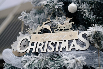 Elegant wooden "Merry Christmas" ornament with Santa sleigh and reindeer hanging on snowy flocked Christmas tree. Festive holiday decoration in rustic gold and white tones for winter season