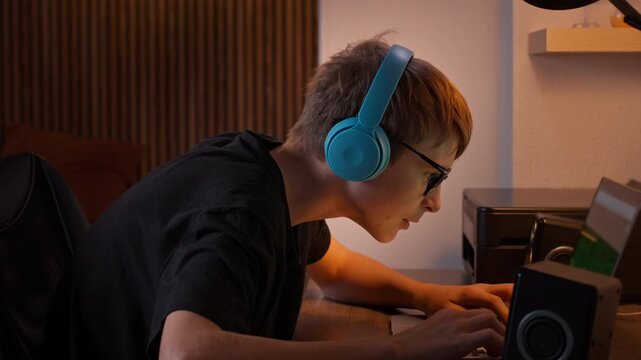 Teenage boy with headphones and glasses intensely play video game on laptop, sit at desk in dark room at night with screen's glow illuminating his focused face